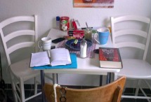 2 weisse Stühle und ein brauner Stuhl um eine Tisch der an der Wand steht. auf dem Tisch stehe Kaffeetassen Zucker, Teller Gebäck. Im Vordergrund liegen Bücher